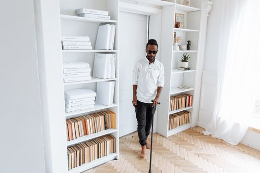 A visually impaired man using a walking stick in a bright room filled with books and shelves.