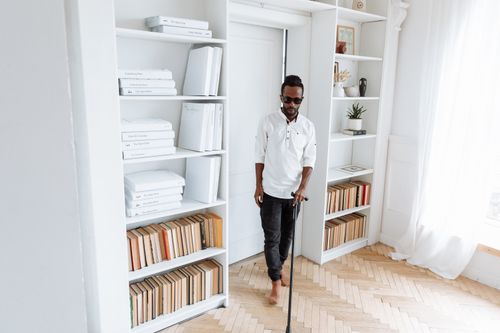 Free A visually impaired man using a walking stick in a bright room filled with books and shelves. Stock Photo