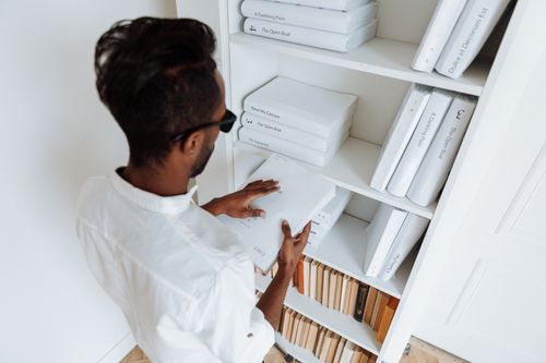 Free A man wearing sunglasses arranges white-covered books on a shelf indoors. Stock Photo