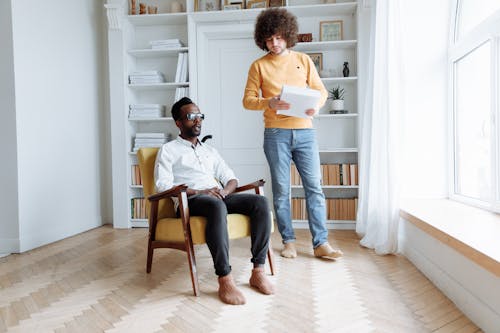 Man in White Long Sleeves Sitting on a Chair Near a Man Holding a Book