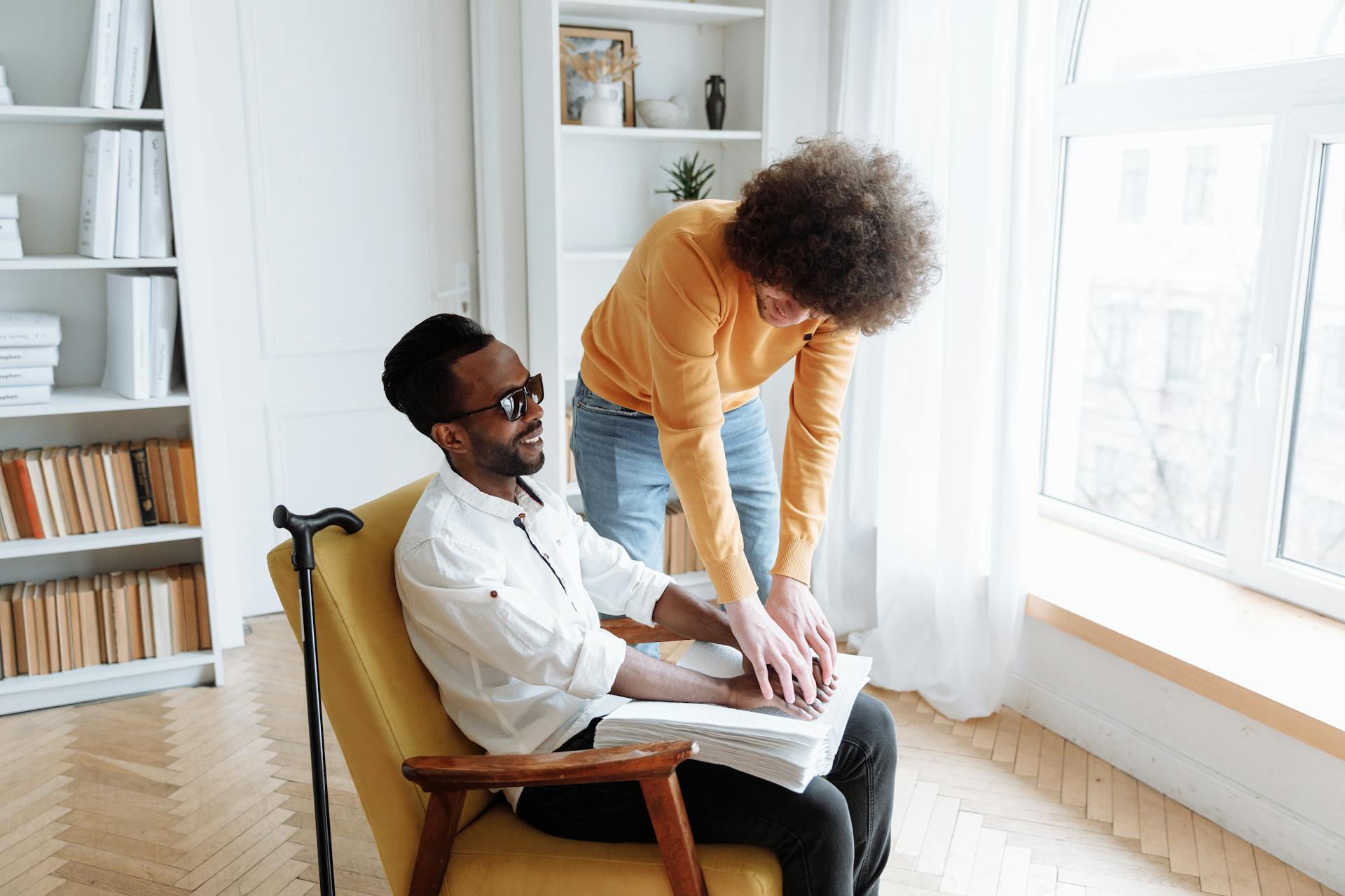 Young Man in Orange Long Sleeve Shirt Assisting a Blind Man Sitting on a Chair Touching a Book in Braille