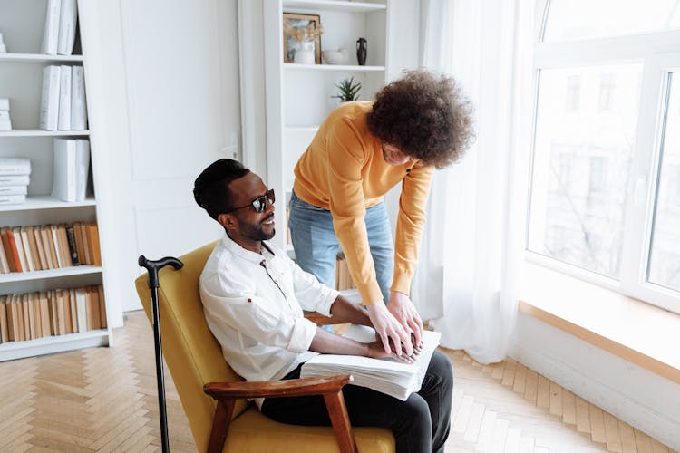 Young Man In Orange Long Sleeve Shirt Assisting A Blind Man Sitting On A Chair Touching A Book In Braille