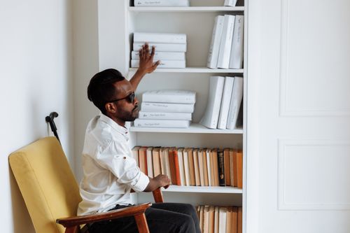 Free African American man using sensory perception to explore a bookshelf indoors, embracing independence. Stock Photo