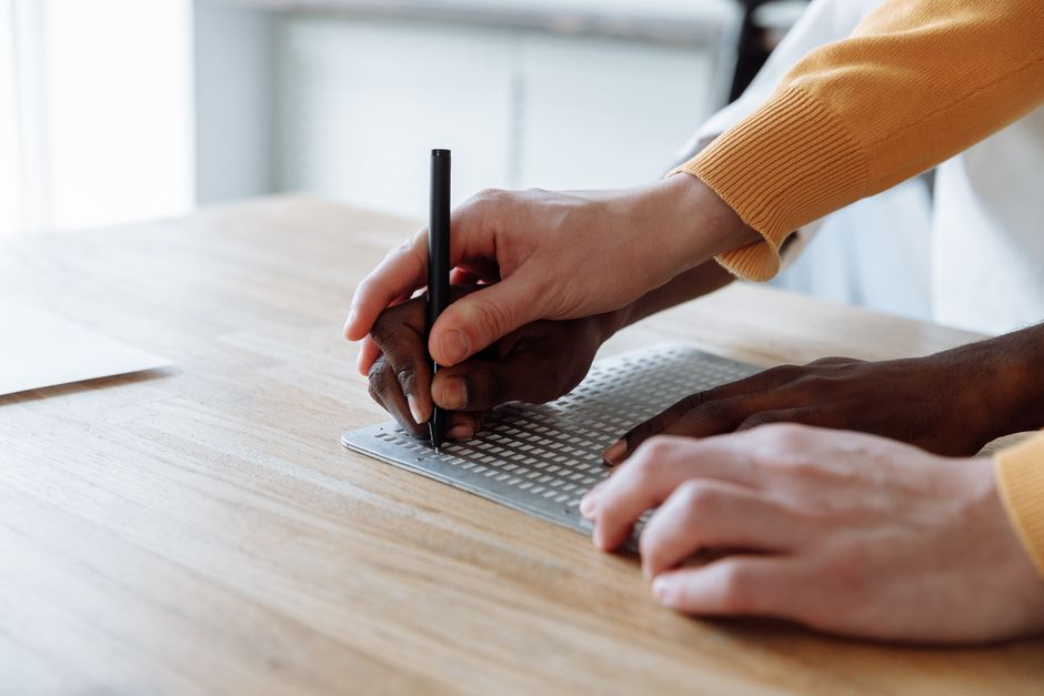 Hands skillfully guiding a student in learning braille on an embossed board.