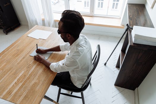 An African man in glasses writing Braille at a wooden desk in a bright indoor setting.
