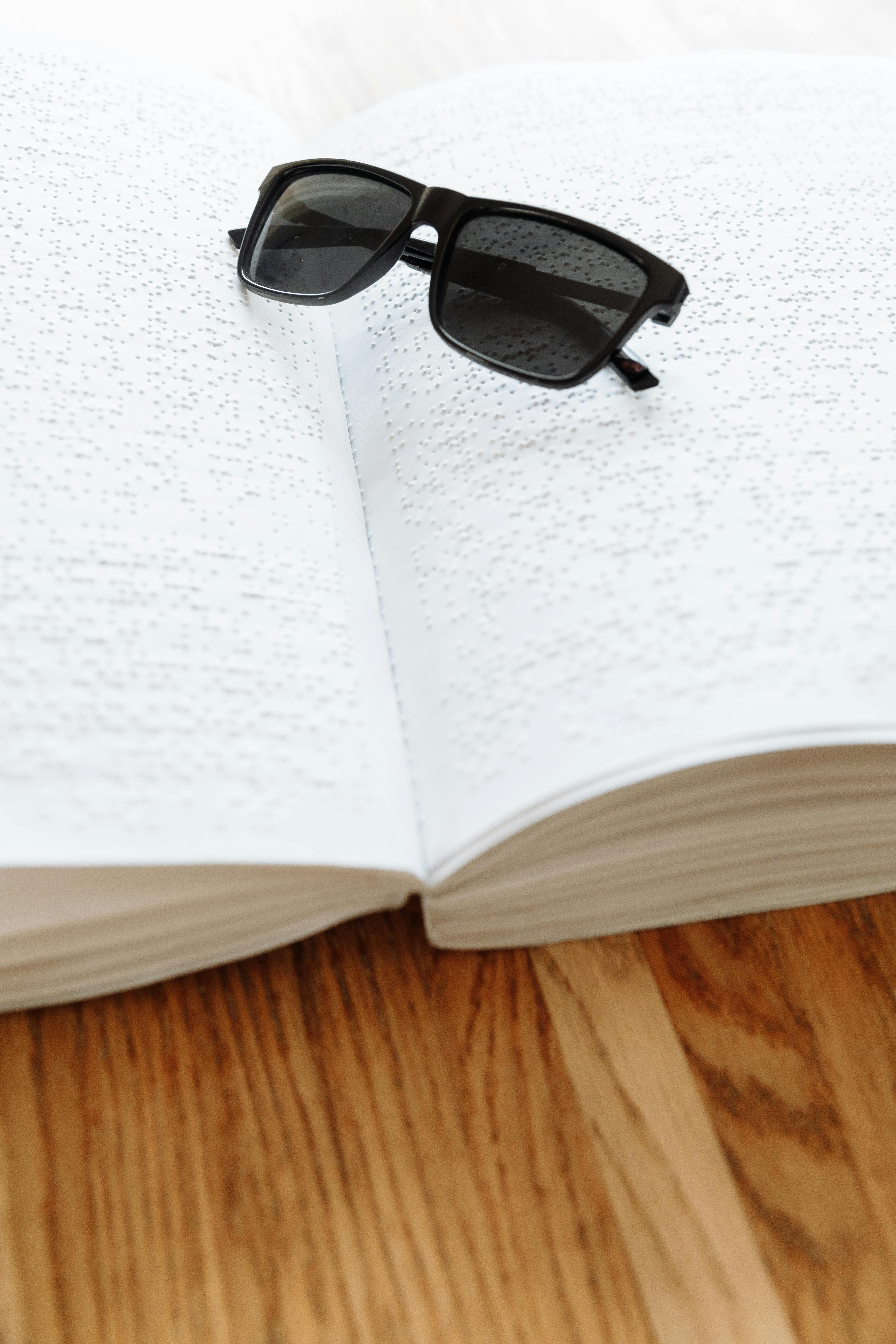 Sunglasses placed on an open Braille book on a wooden table, emphasizing accessibility.