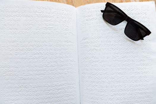 Close-up of an open Braille book with sunglasses on a wooden table.