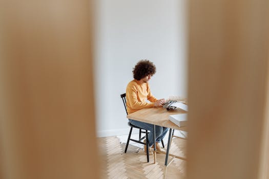 Focused man with curly hair types on laptop while seated indoors in casual attire.