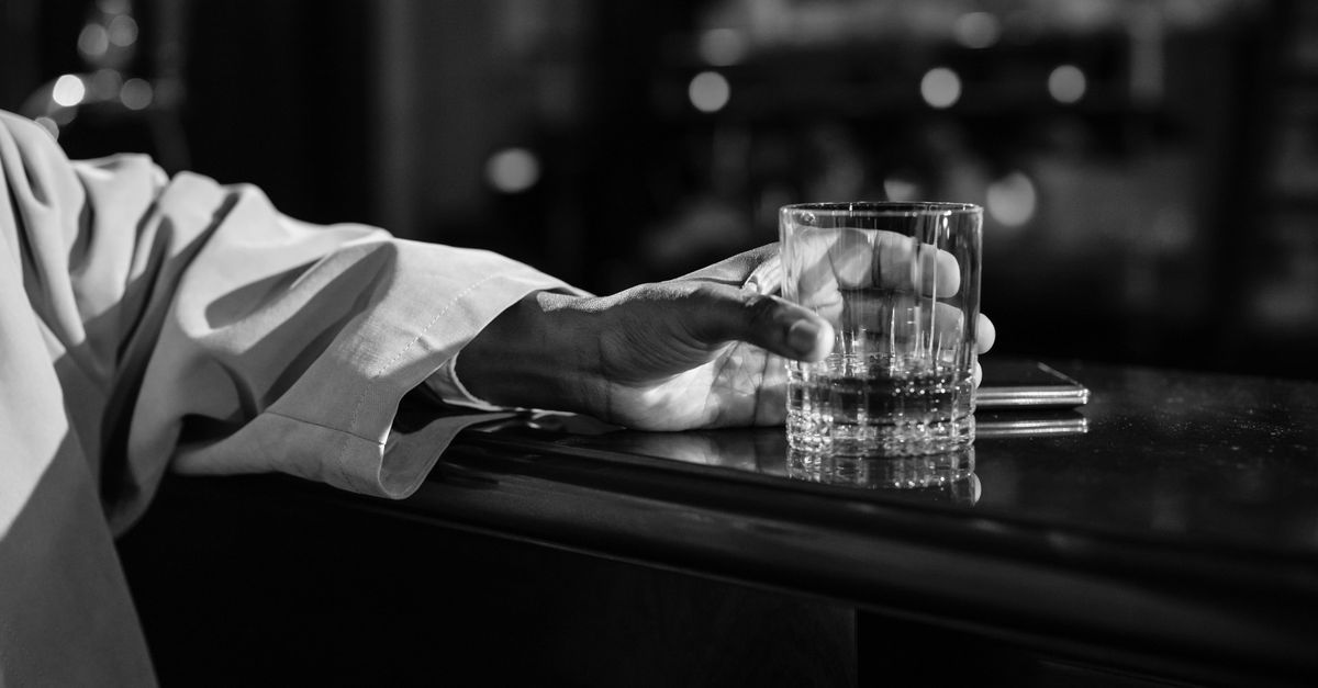Moody black and white image of a hand holding a whiskey glass on a bar counter.