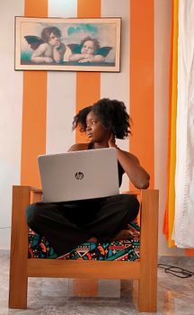 Young black woman sitting on a chair, using a laptop in a vibrant living room.