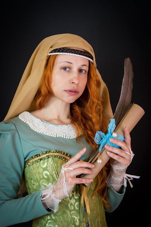 Free Serious young female in creative costume with long ginger hair holding scroll and feather while looking at camera on black background Stock Photo
