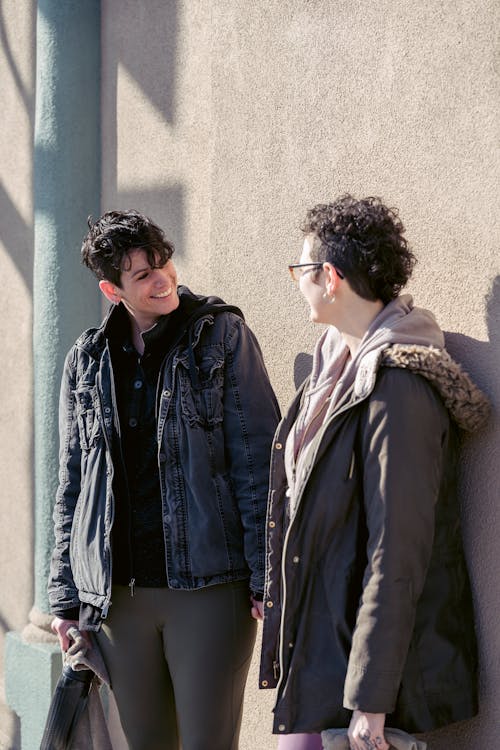 Lesbian couple in warm clothes holding hands and looking at each other in city street near building wall in sunny day