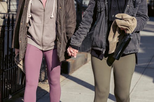 Crop anonymous lesbian couple in warm clothes holding hands and strolling together on sunny autumn street