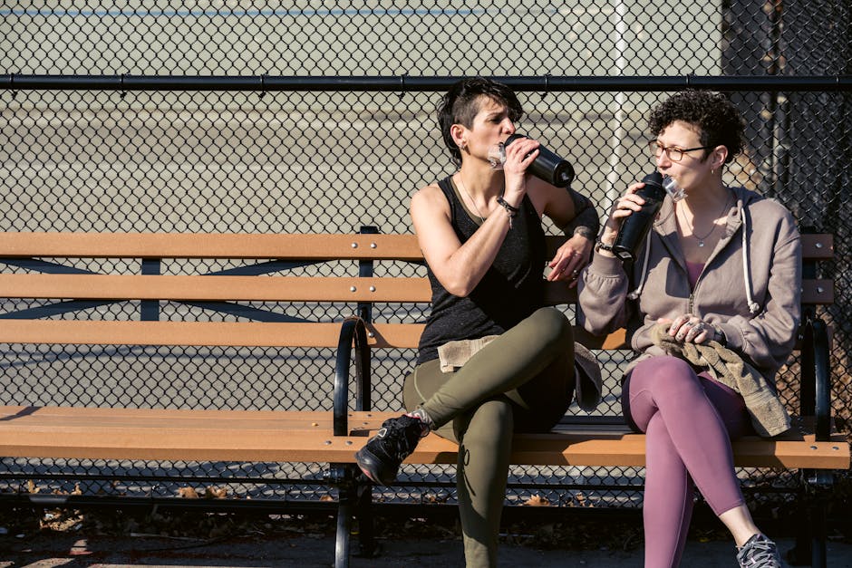 Active female friends in sportswear sitting on bench and drinking water from bottles while resting after workout in park