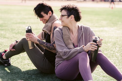 Free Couple of thirsty athletic girlfriends talking and smiling while drinking water and sitting on grass on stadium in back lit in daylight Stock Photo