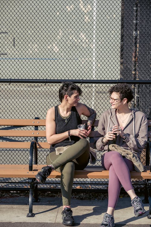 Free Full body of sportswomen chatting and smiling while relaxing on bench with bottles of water in hands in daylight Stock Photo
