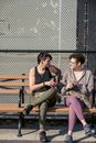 Full body of sportswomen chatting and smiling while relaxing on bench with bottles of water in hands in daylight