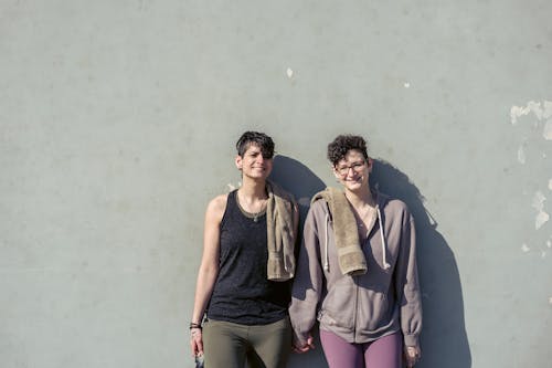 Free Cheerful female couple of athletes in love wearing sportswear looking at camera and smiling while holding hands and standing against gray wall in daytime Stock Photo