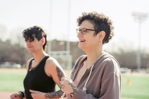 Free Side view of couple of girlfriends in activewear jogging while cheerful female athlete laughing with androgynous female at stadium in back lit in daytime Stock Photo