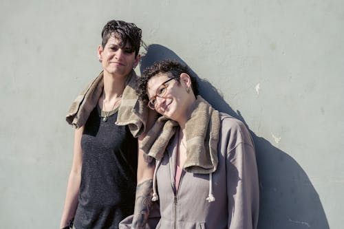 Free Happy gay couple of female athlete leaning on shoulder of androgynous girlfriend and relaxing while smiling and standing together against gray wall in daylight Stock Photo