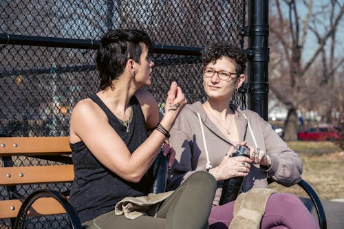 Young females in sportswear after workout in park resting and talking to each other while sitting on bench