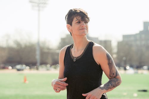 Free Positive young androgynous female athlete with short hair and tattoo on arms smiling while running in city park during outdoor workout on sunny morning Stock Photo