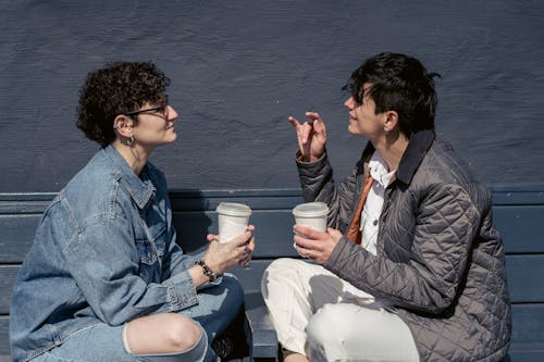 Free Side view of young stylish ladies with short dark hairs in casual outfits sitting on wooden bench with crossed legs and chatting while drinking takeaway coffee during weekend on street Stock Photo