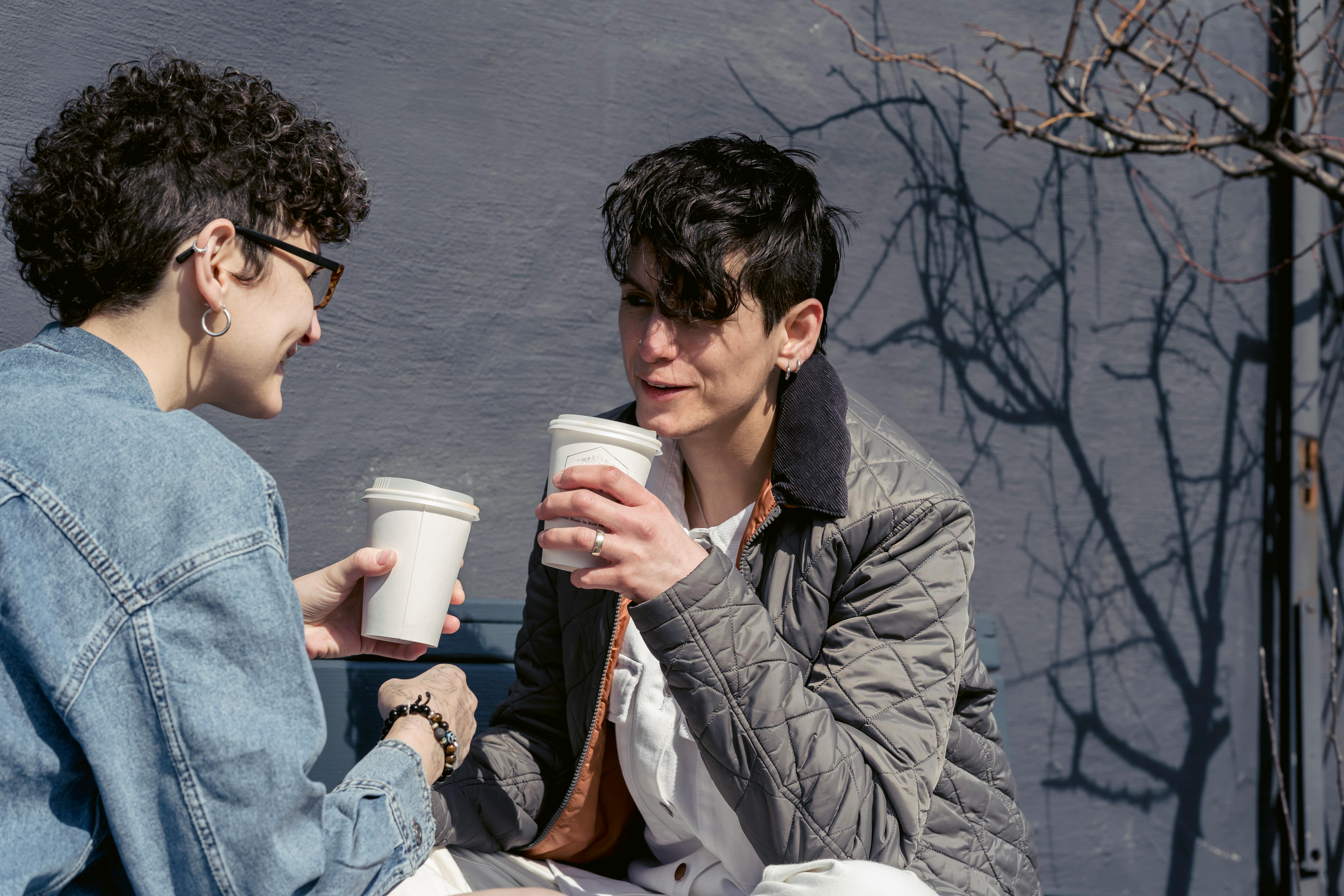 joyful young female friends drinking coffee and talking on bench in morning