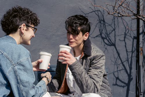 Cheerful young trendy female best friends with short hair in casual clothes smiling while drinking takeaway coffee and gossiping on bench on sunny day