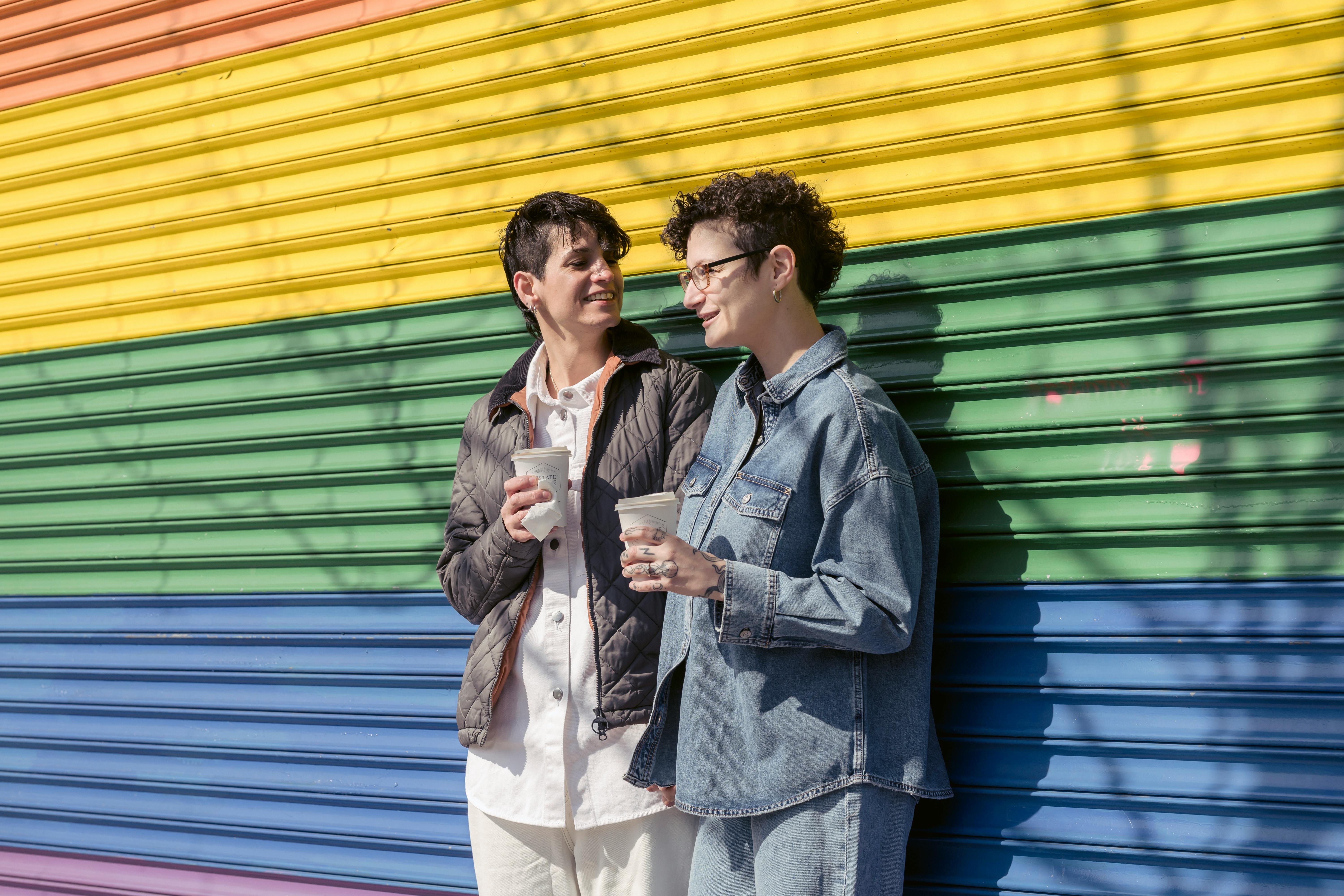 stylish lesbian couple with takeaway coffee cups standing near rainbow colored wall