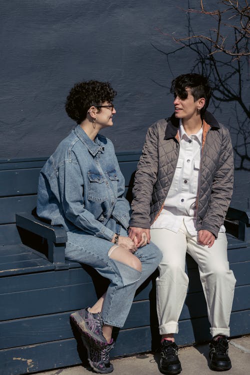 Free Full body of positive young lesbian couple with short dark hair in stylish clothes holding hands and speaking with each other while sitting on bench on street on sunny day Stock Photo