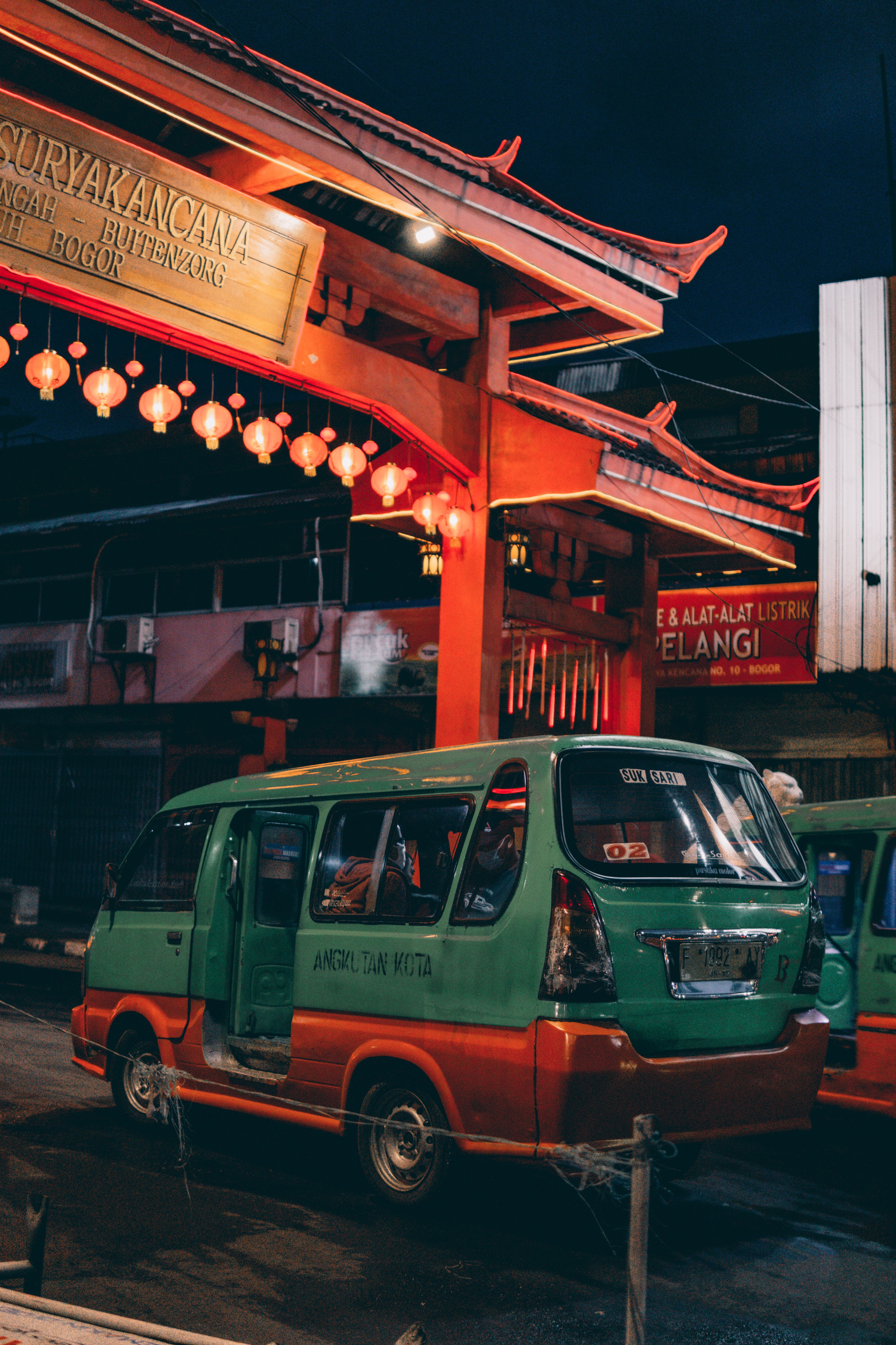Lively night shot of an angkot public transport vehicle under vibrant red Chinese gate in Bogor, Indonesia.