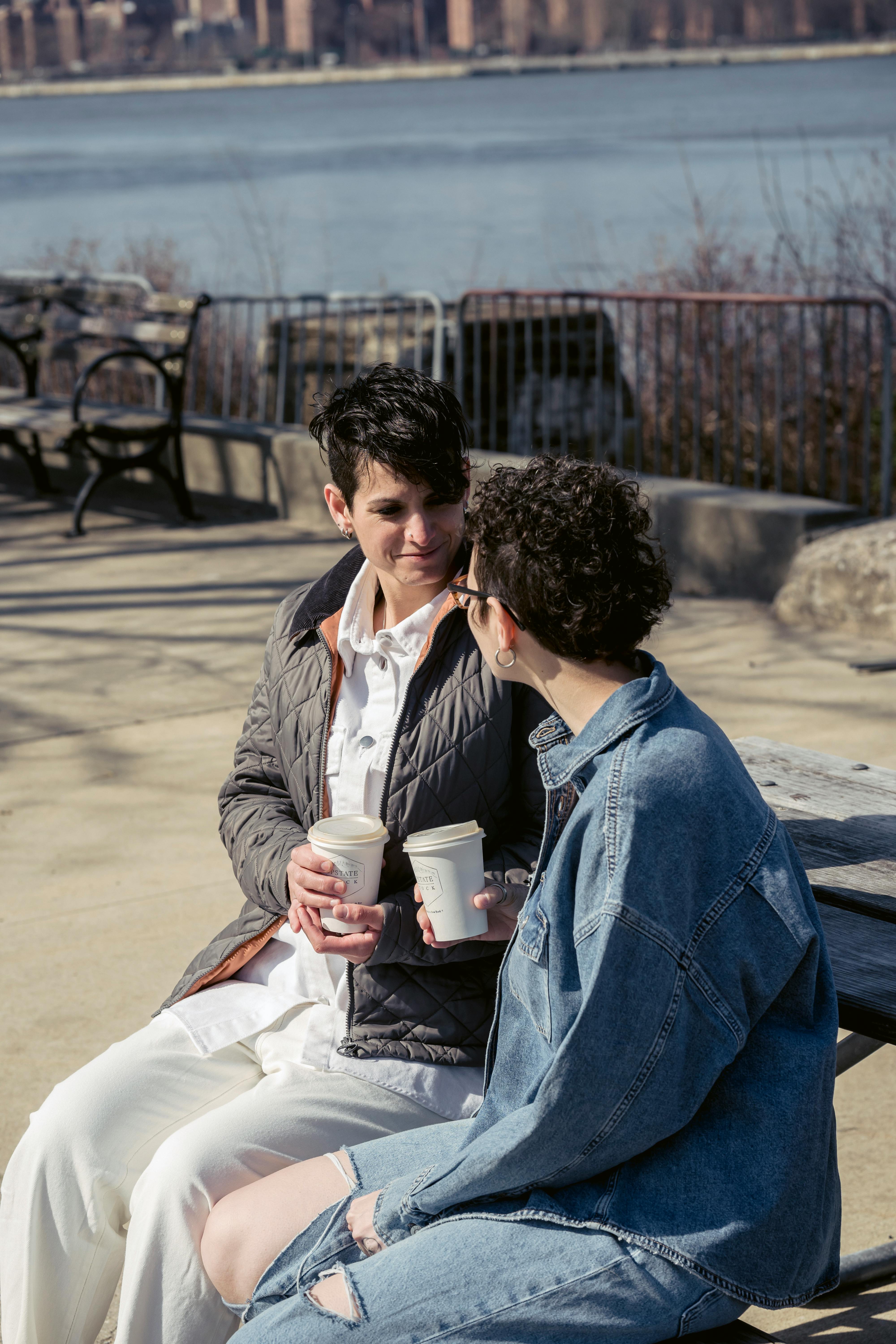 happy young ladies drinking coffee on bench on river embankment