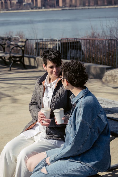 Free Positive young trendy female friends with short dark hair in casual clothes chatting on bench on river embankment and drinking takeaway coffee during break in sunlight Stock Photo