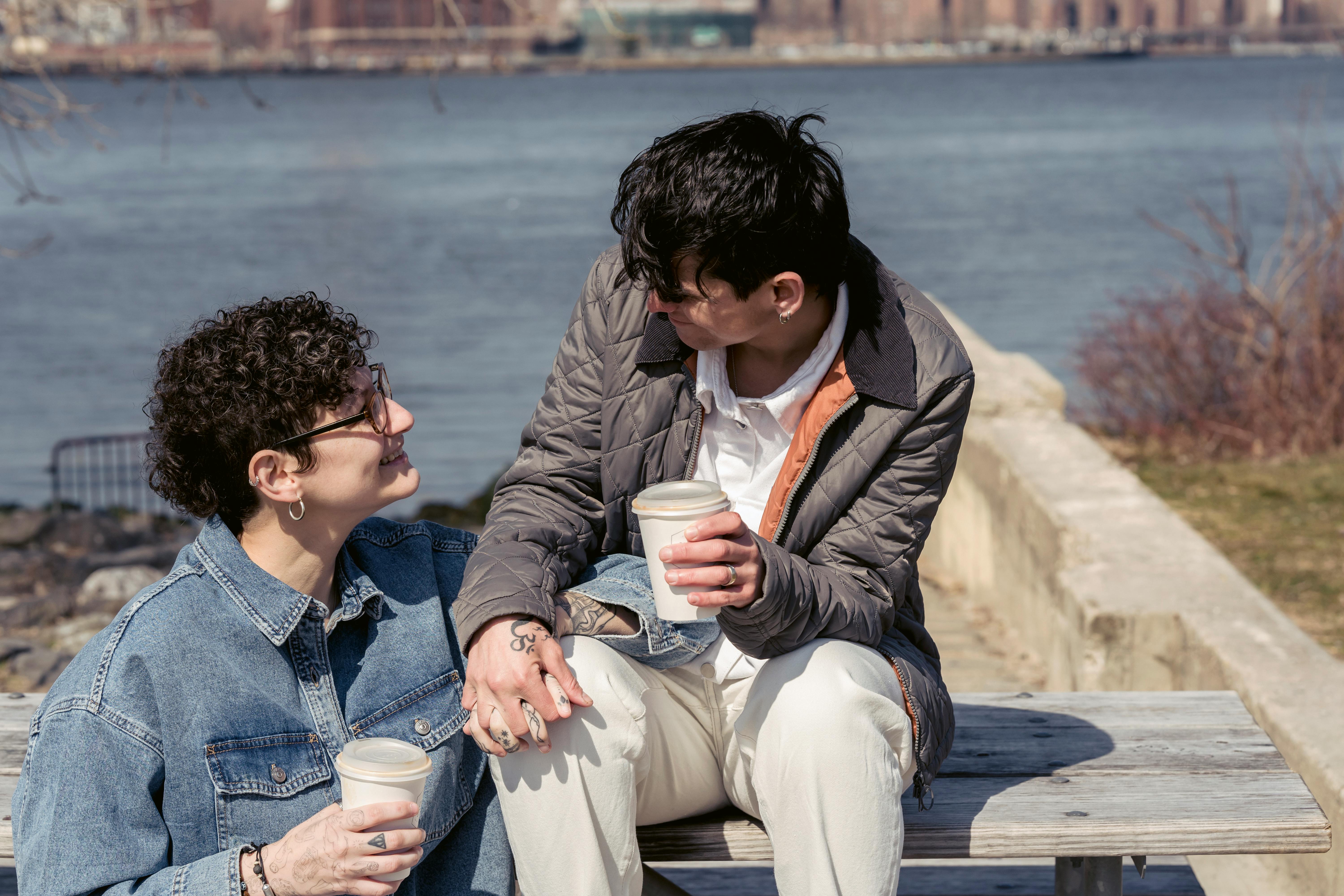 happy young homosexual ladies smiling and holding hands during coffee break on river embankment