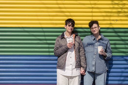 Trendy young lesbian couple with similar stylish haircut in casual clothes holding hands and drinking takeaway coffee near pride rainbow wall on sunny day on street