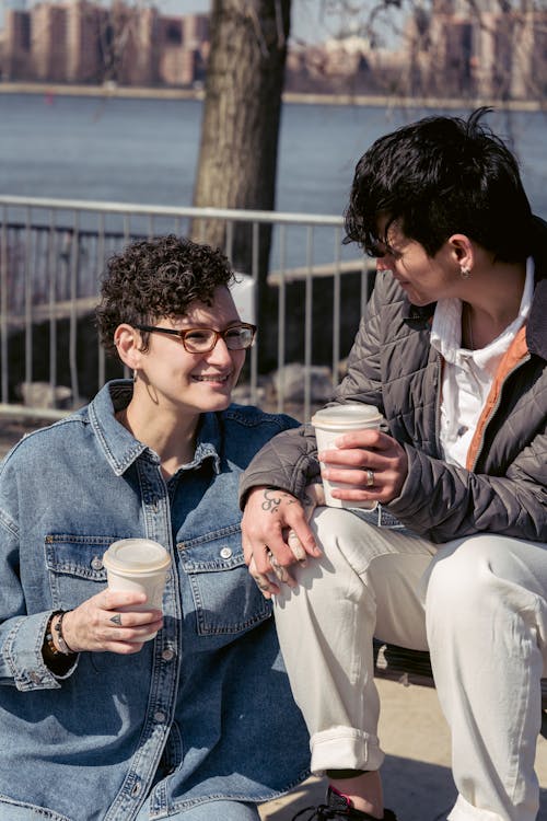 Positive trendy young lesbian couple holding hands and smiling while chilling on bench at riverside during coffee break in city