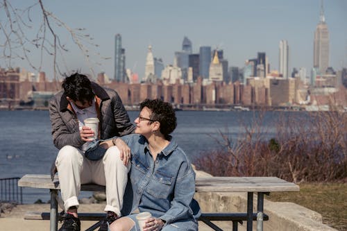 Free Trendy young lesbian couple smiling and drinking takeaway coffee while chilling on wooden bench at riverside in modern city Stock Photo