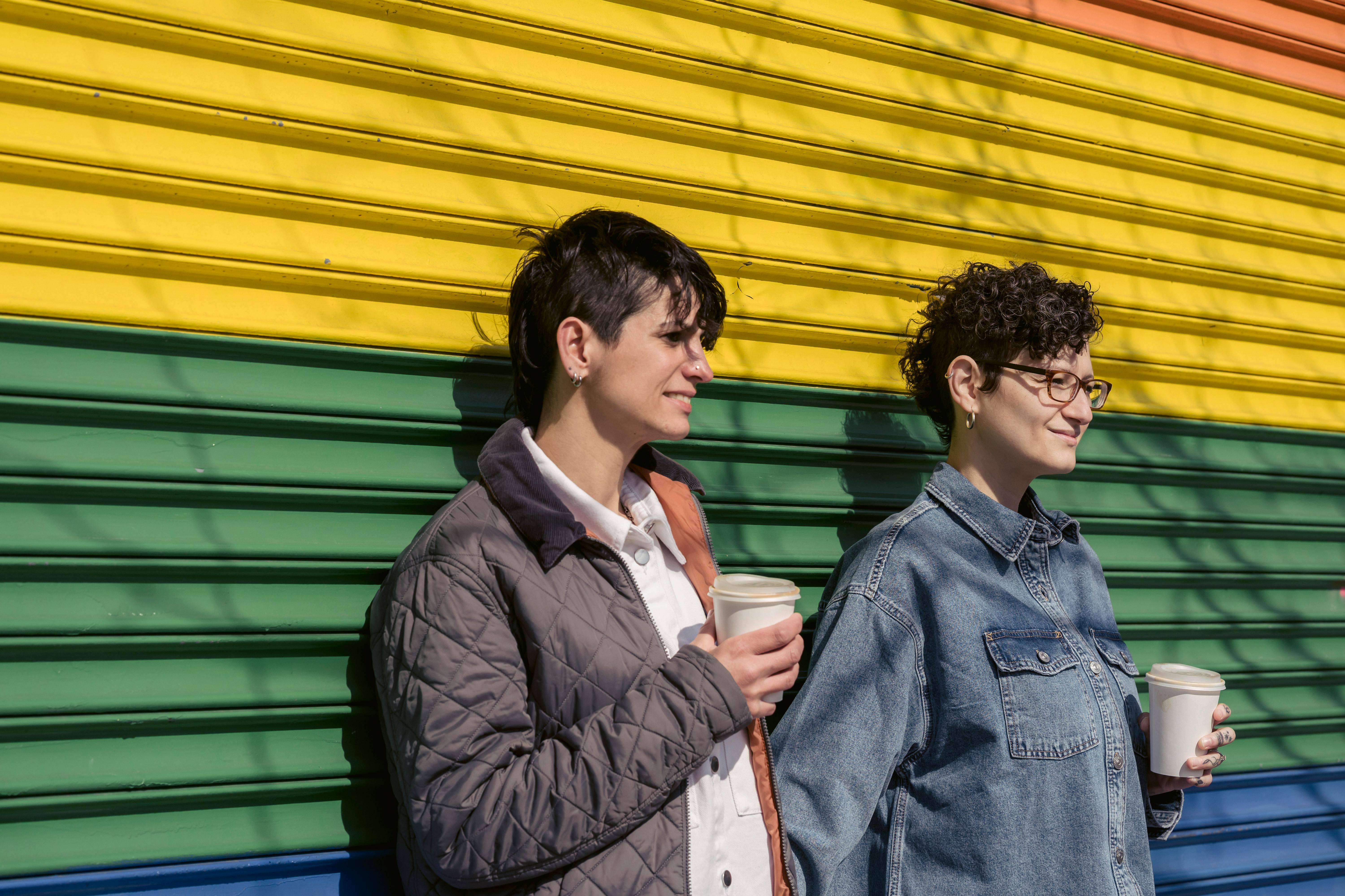 smiling homosexual girlfriends standing near rainbow wall with cups of takeaway beverages