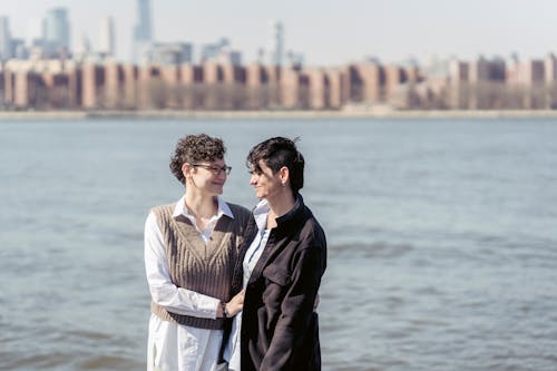 Positive women with dark short hair smiling and supporting each other on blurred background of river with embankment with buildings