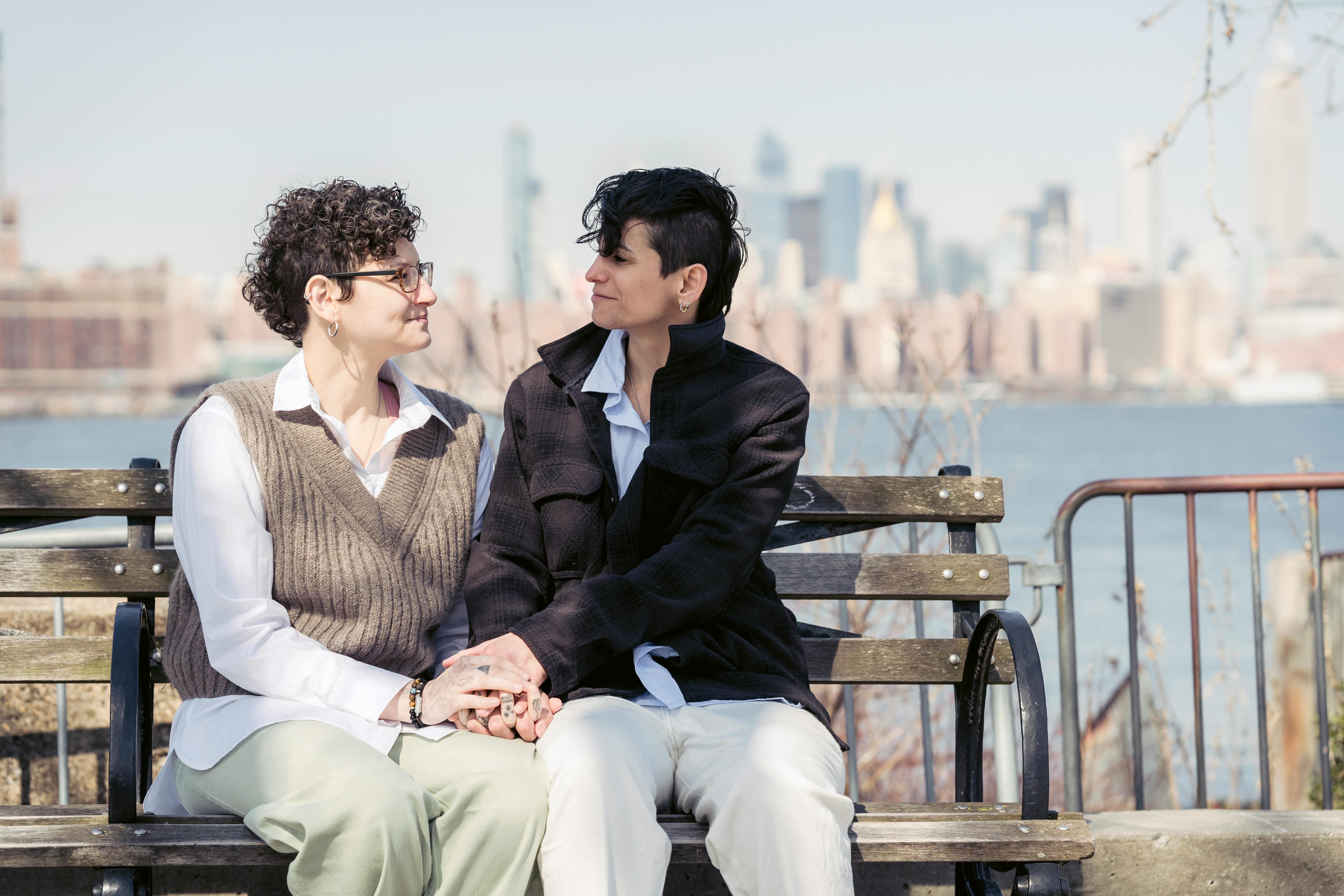 lesbian couple sitting on bench and holding hands near river in city