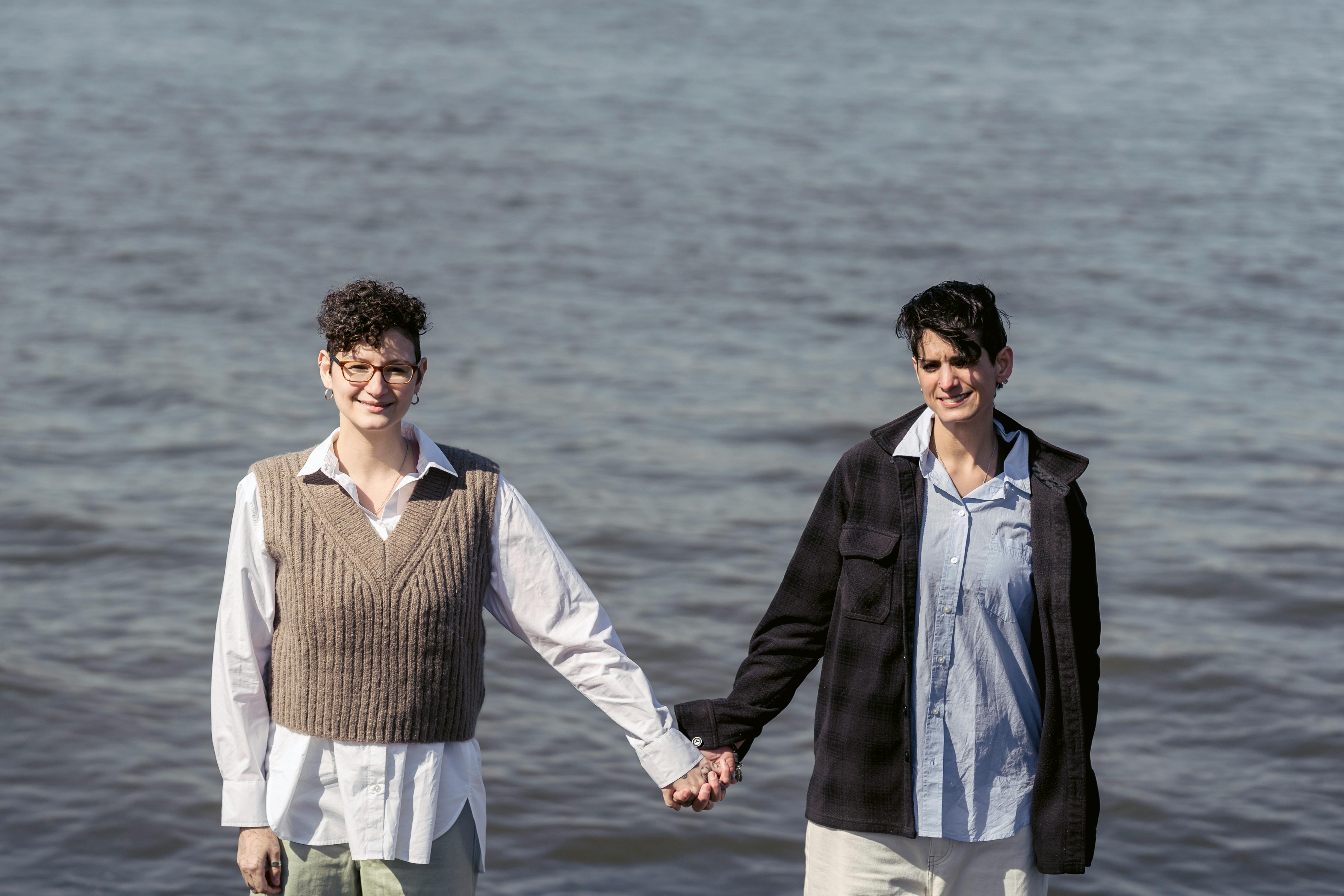 positive women in casual clothes holding hands against river