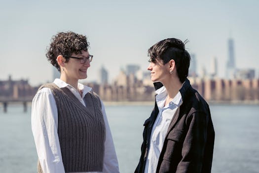 Side view of cheerful woman standing and talking with androgynous female on embankment near river against cityscape in sunny day