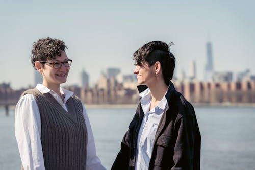 Free Positive lesbian couple holding hands and smiling while chilling at riverside in city Stock Photo