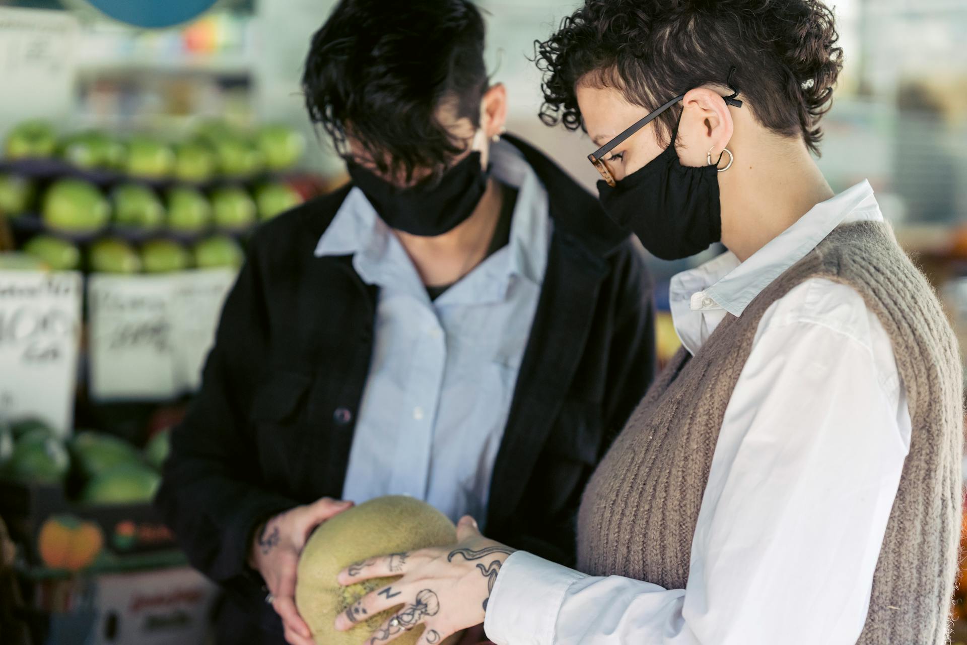 Crop unrecognizable female friends with short dark hair in casual clothes and protective masks examining melon while purchasing products in grocery market