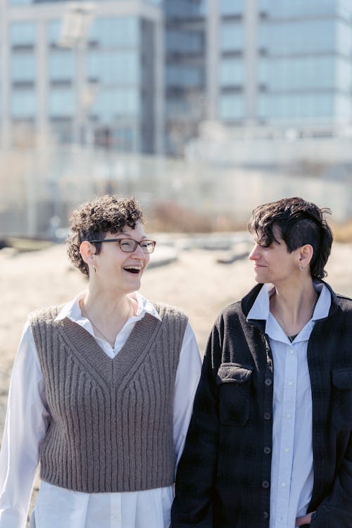 Joyful young female best friends with short dark hair in casual clothes laughing and looking at each other while strolling together in city on sunny day