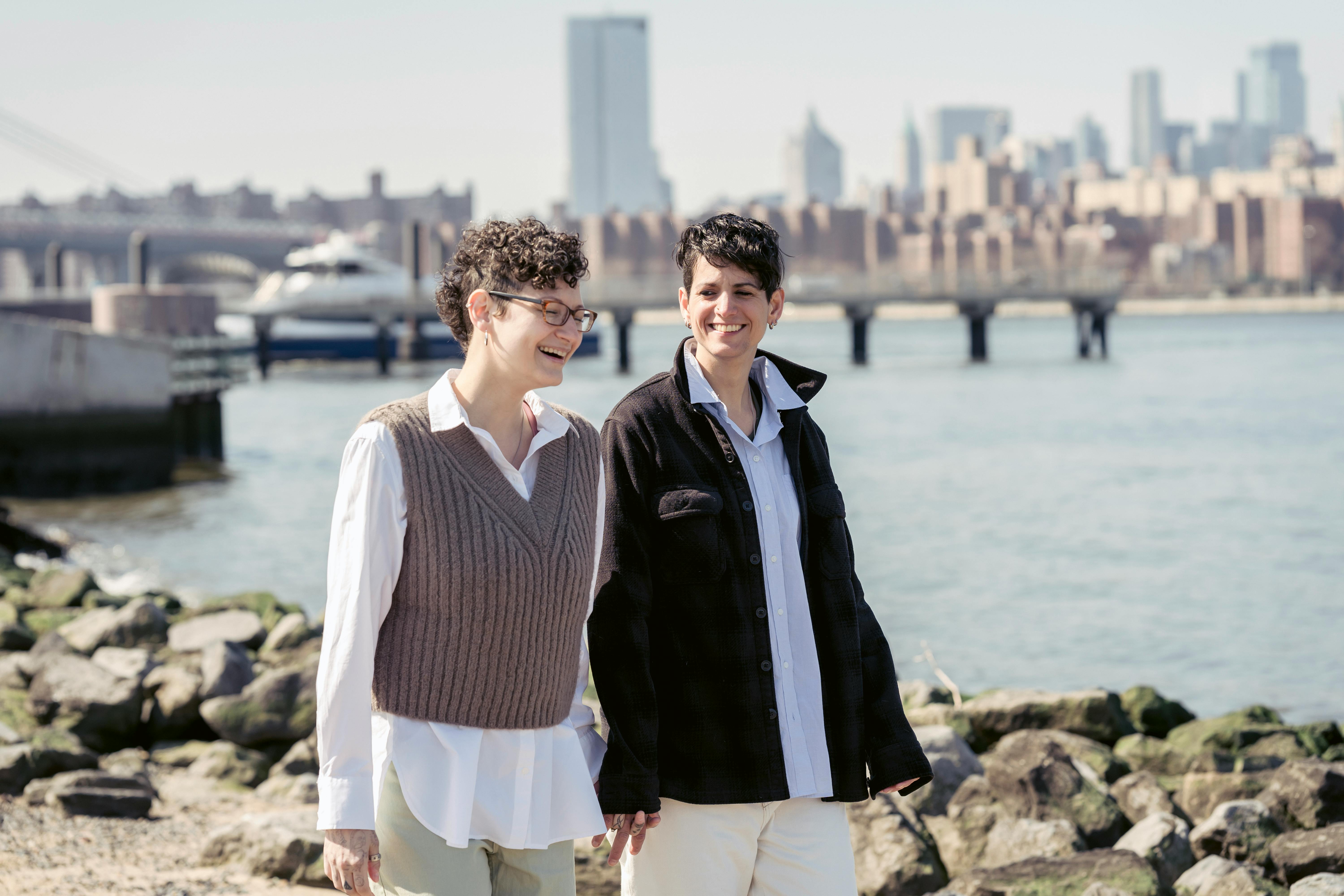 joyful young lesbians holding hands and walking near river in city