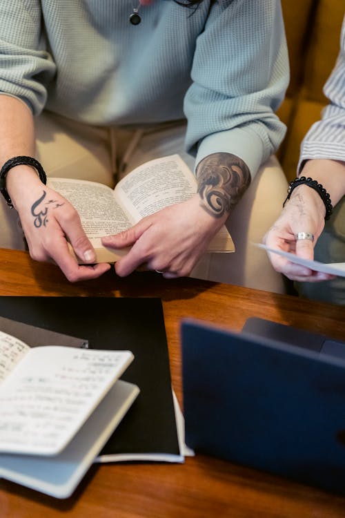 Free From above of crop unrecognizable tattooed person sitting on sofa and reading interesting book near friend working remotely on laptop Stock Photo