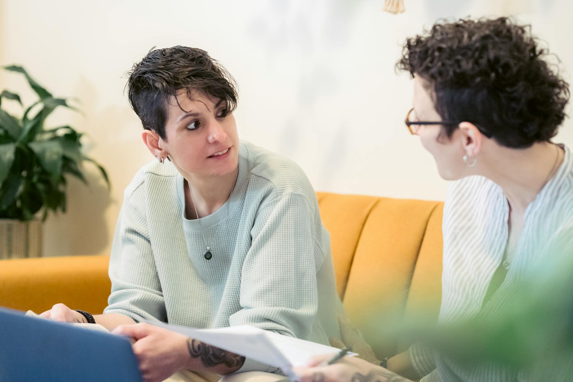 Positive young female remote specialists with short dark hair in casual clothes sitting on sofa and chatting while working online on laptop and reading documents