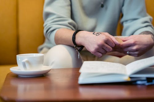 Free Crop anonymous tattooed person in casual clothes reading interesting book while sitting on comfortable sofa with cup of coffee Stock Photo